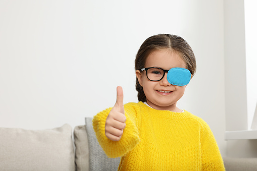 Happy girl with nozzle on glasses for treatment of strabismus showing thumbs up in room. Space for text