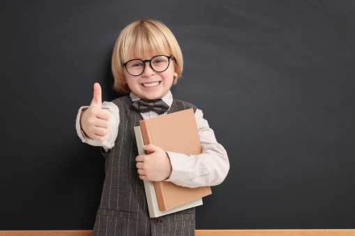 Happy little school child with notebooks showing thumbs up near chalkboard. Space for text