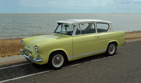 Felixstowe, Suffolk, England - August 29, 2015: Classic Yellow Ford Anglia motor car on seafront  promenade beach and sea in background.
