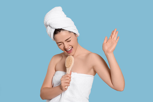 Emotional woman in towels singing into brush on light blue background. Spa treatment