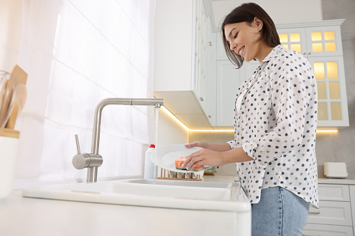 Happy young woman washing plate above sink in modern kitchen