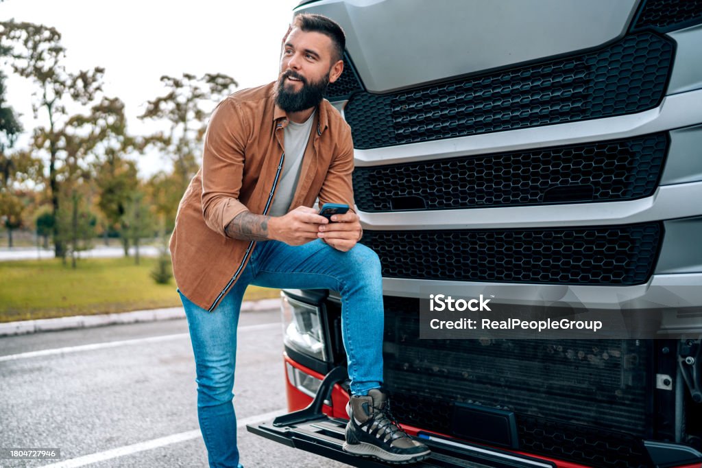 Truck driver sends text in front of his vehicle A truck driver is seen standing in front of his truck, focused on sending a text message on his mobile phone, managing communication amidst his travels Mobile Phone Stock Photo