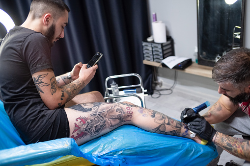 Tattooist painting tattoo on a young man leg in a tattoo studio while the customer using cellphone for leisure.