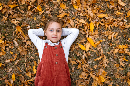 Portrait of cute little girl, with her arms behind head, lying on the ground in the autumn public park, autumn leaves around her.