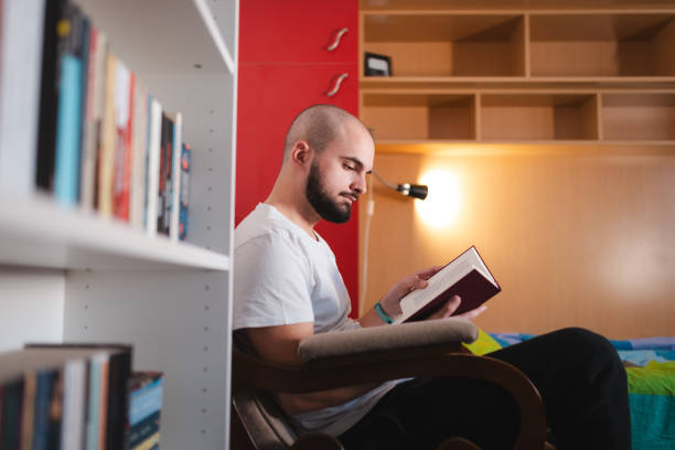 Young man reading a book at home. He is relaxing, sitting by a bookshelf.