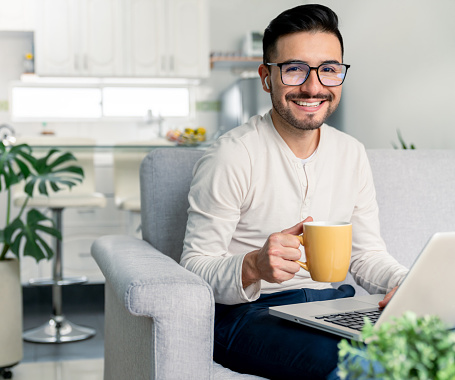 Happy Latin American man working at home and drinking a cup of coffee while looking at the camera smiling