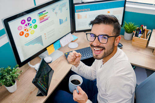 Excited Latin American man working at home on his computer and celebrating an achievement while looking at the camera smiling