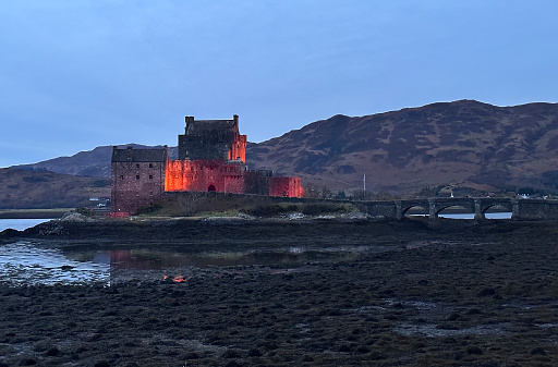 Dornie, Scotland, UK - November 17th, 2023: A low tide view of Loch Duich in the evening with Eilean Donan Castle illuminated.
