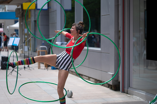 A street entertainer is showing off on the sidewalk by spinning plastic hoops around her arms and legs on a hot day in the city