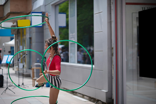 Wearing a stripy costume and standing on one leg, this circus acrobat performs tricks, spinning four hoops around her arms and legs outside on the sidewalk on a sunny day