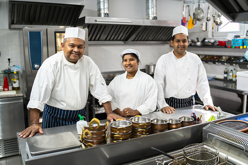 A family of chefs, father son and wife are standing in an Indian restaurant kitchen, leaning on the kitchen counter as they smile and look at the camera