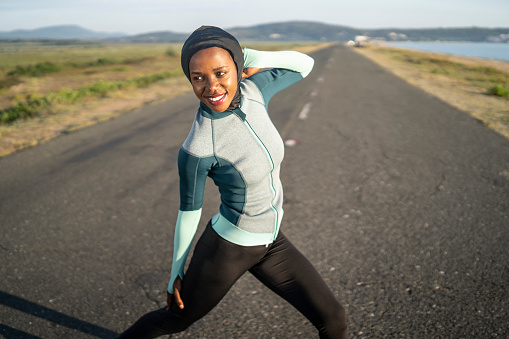 A woman wearing jogging clothing is stretching her arms and legs for a warm up exercise before starting her road running along a long straight country road beside a narrow beach