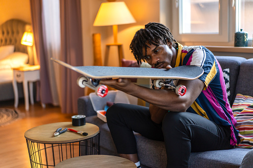 A multiracial man is looking at the bottom of a skateboard to check the wheels, holding it up to see clearly from a couch in a modern living room