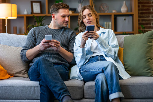 Young couple sit on sofa holding smart phone. Wife show explains to husband new mobile apps, comparing applications enjoy modern tech usage on weekend at home