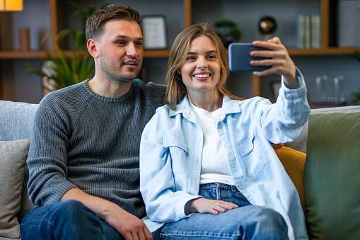 Happy cheerful young couple having online webcam chat with via video call on mobile phone, while sitting together in the cozy apartment. Concept of household life