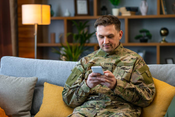 istock Soldier in uniform sitting on the sofa and using cellphone texting chatting with friend. 1804866648