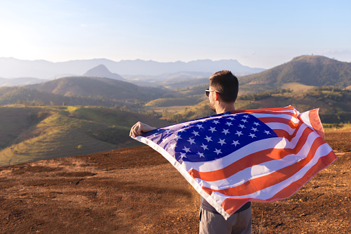 Man with the American flag unfurled atop a mountain against a backdrop of mountainous landscapes. The pride of being American. Ideal for campaigns that celebrate unity during sporting events or on the 4th of July.