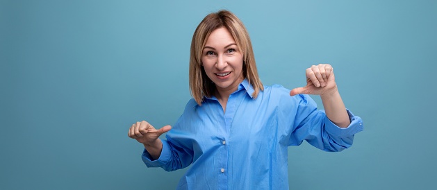 confident blond young millennial in casual shirt shows hands super at herself on blue background with copy space.