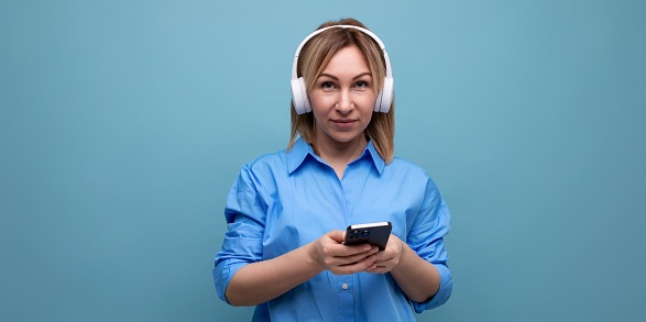close-up of a millennial woman in a casual shirt with big white headphones with a smartphone in her hands on a blue isolated background with copy space.