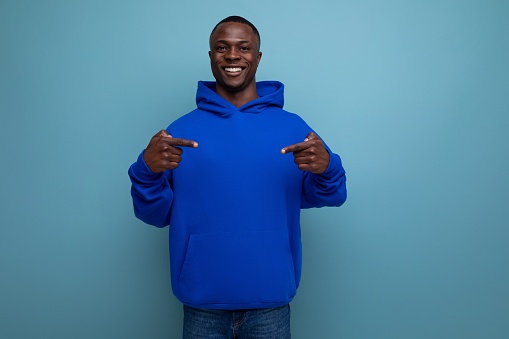 confident dark-skinned young american man in a blue sweatshirt shows his hand at an advertisement on a studio background with copy space.