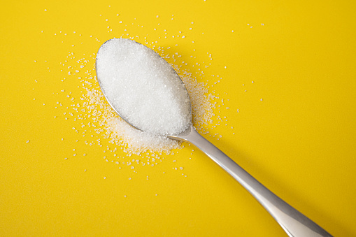 A silver stainless steel spoon filled with sugar or salt that is overflowing on to a yellow backdrop