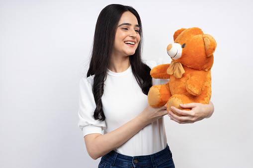 Smiling little Asian girl raised up stuffed teddy bear while sitting on the bed at home.