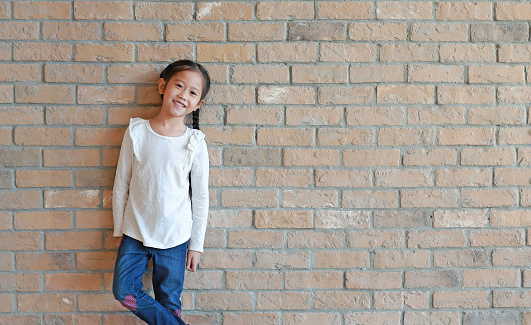 Portrait of happy asian little girl with pigtail hair in jeans and a white shirt standing at brick wall background with copy space.