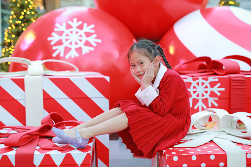 Cute little child girl in red dress poses sitting on big gift in winter season and happy new year festival against christmas background.