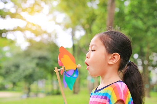 Adorable little Asian kid girl blowing wind turbine in the summer garden.