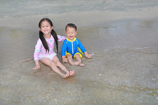 Happy little Asian sister and her little brother in swimming suit sitting and playing sea waves on the beach.