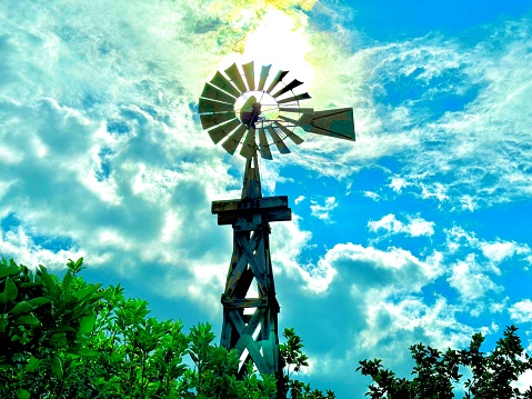 A wooden farm windmill sits still. Behind the windmill is a bright yellow sun casting a brilliant aura. The sky is bright blue with fluffy clouds. Lush green trees boarder the bottom of the image and frame the windmill