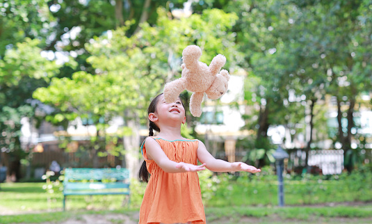 Portrait of happy little Asian child in green garden with throwing up teddy bear doll floating on air. Smiling kid girl playing in summer park.