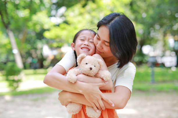 portrait d’une mère asiatique heureuse câlinant sa fille et étreignant une poupée d’ours en peluche dans le jardin. maman et fille enfant avec l’amour et le concept de relation. - enfold photos et images de collection