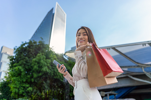 Young Asian busineewoman in Casual Business Outfit Enjoying Shopping Spree, Juggling Smartphone and Shopping Bags in a Central Business district against the backdrop of corporate buildings. Business on the Move. Connected Mobility.