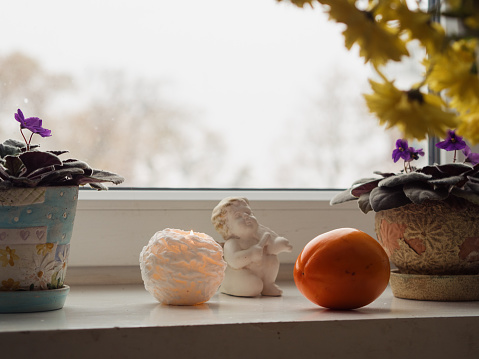 In this horizontal close-up, a serene white windowsill hosts a picturesque arrangement. Two pots of blooming violets stand gracefully, flanked by a vibrant orange persimmon, casting a burst of color. A softly lit candle adds a warm glow to the scene, illuminating the delicate details of a statue of an angel. In the foreground, yellow chrysanthemums introduce a touch of elegance, creating a harmonious composition that seamlessly blends natural beauty with subtle, ambient light.