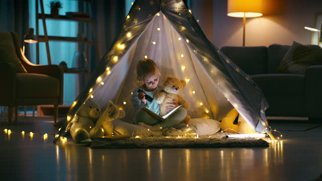 Story, flashlight and teddy bear with a girl in a tent in the bedroom of her home to camp at night. Kids, book or storytelling with a young child reading to her stuffed animal to relax at bedtime