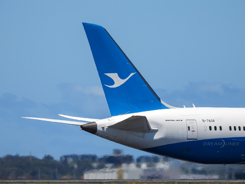 The tail and fuselage of a Xiamen Air Boeing B787-9 plane, registration B-7838, taxiing to the southern end of Sydney Kingsford-Smith Airport in preparation for departure as flight MF802 to Xiamen, China. This image was taken from Botany Bay, Kyeemagh on a sunny afternoon on 18 November 2023.