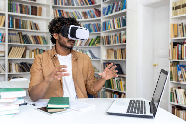 istock Young hispanic male student sitting at a desk in a university library, studying online on a laptop wearing a virtual mask. Gesturing and waving his hands 1805415192