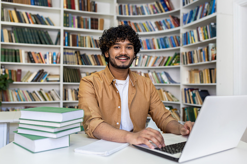 Portrait of a smiling young Indian man studying and working in a library at a laptop. Sitting at a table with books and looking at the camera.