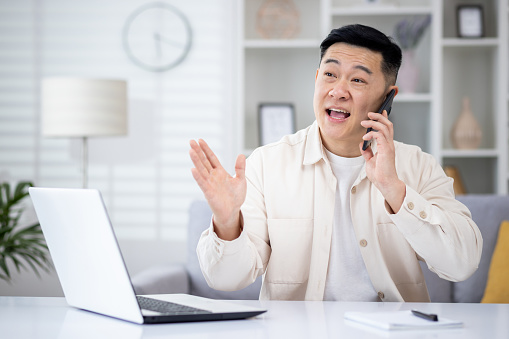 Mature asian man talking on the phone at home, man working in home office sitting at desk in home office, happy and smiling, consulting clients remotely.