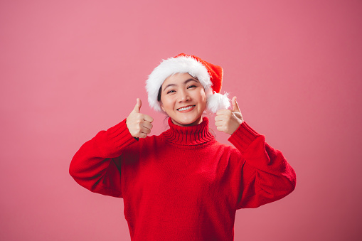 Asian woman wearing Santa hat happy showing thumb up, looking at camera, standing isolated on pink background.