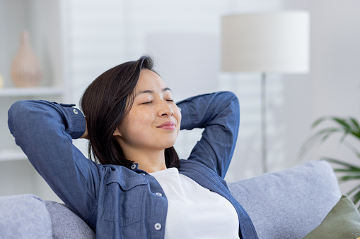 Young beautiful Asian woman relaxing at home close-up on sofa in living room, hands behind head with eyes closed dreaming and visualizing future results achievement and plans.