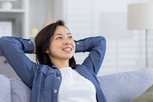 Closeup photo of a young beautiful Asian at home in the living room, woman has her hands behind her head and is resting, breathing fresh air, smiling and looking window, dreaming happy future.