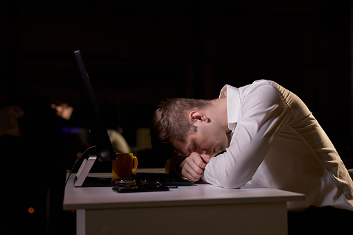 Young caucasian male sleeping at work place in office at night, need more rest, has no more strength to work at late night. side view on exhausted man in formal wear with head bowed on desk
