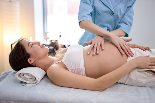 Young pregnant woman receiving tummy massage in spa center. Female patient is receiving treatment by professional therapist with beautiful hands. Body positive, harmony, healthy lifestyle. side view