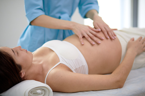 Pregnant young woman with beautiful skin lying on bed having relaxing prenatal massage, physiotherapist doing various techniques for better massaging, focus on brunette mom-to-be