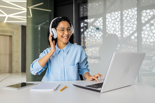 Indian woman in good mood, office worker in headphones, sitting at desk in front of laptop in modern office, Hispanic woman listening to music during work break, talking on video call.