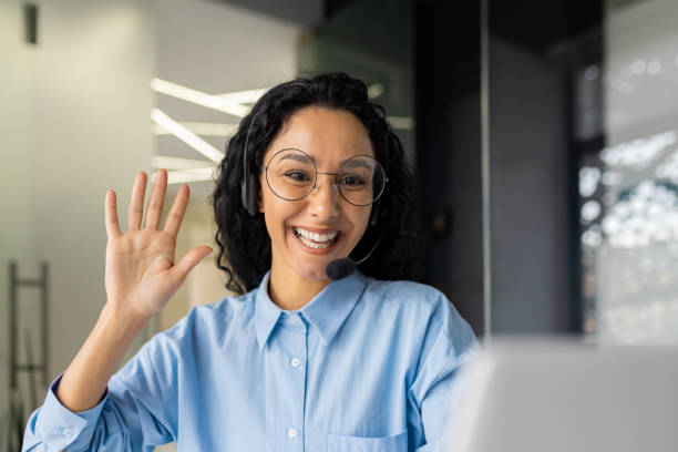 istock Young female online support worker using headset phone for video call, consulting customer consumers remotely, businesswoman working inside office with laptop, waving at camera saying hello 1805564456