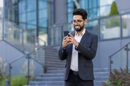 A young businessman in a business suit and glasses is walking around the city outside an office building, the man is holding a phone in his hands, typing a message and using an application.