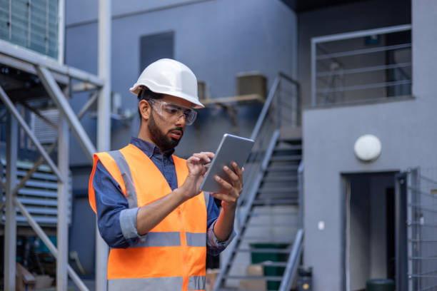 Serious thinking and focused engineer working in factory in hard hat and vest, man using tablet computer reading diagram, and checking equipment, inside industrial factory